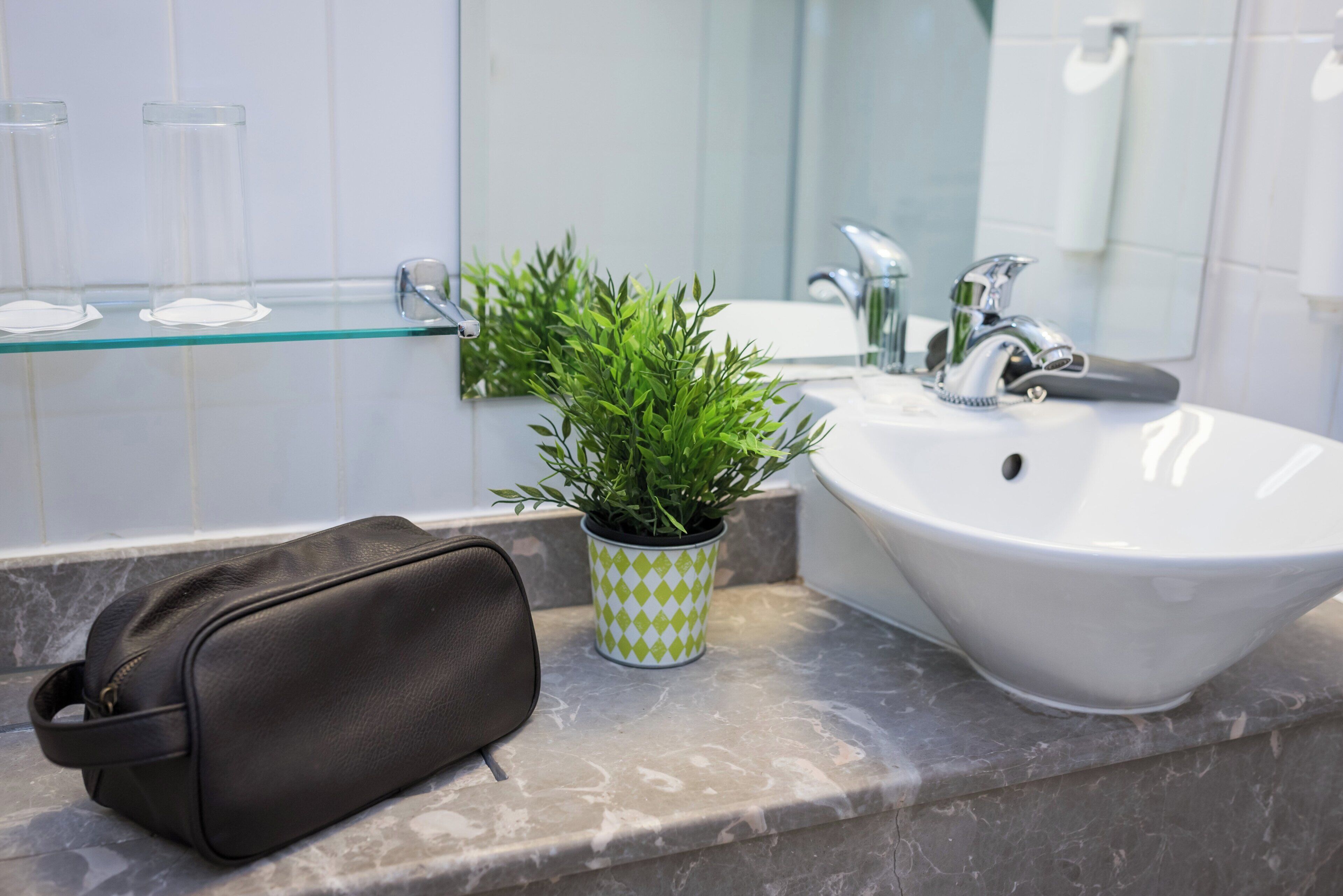 Un baño con encimera de mármol, un lavabo blanco, estante de vidrio, planta en maceta y una bolsa negra.