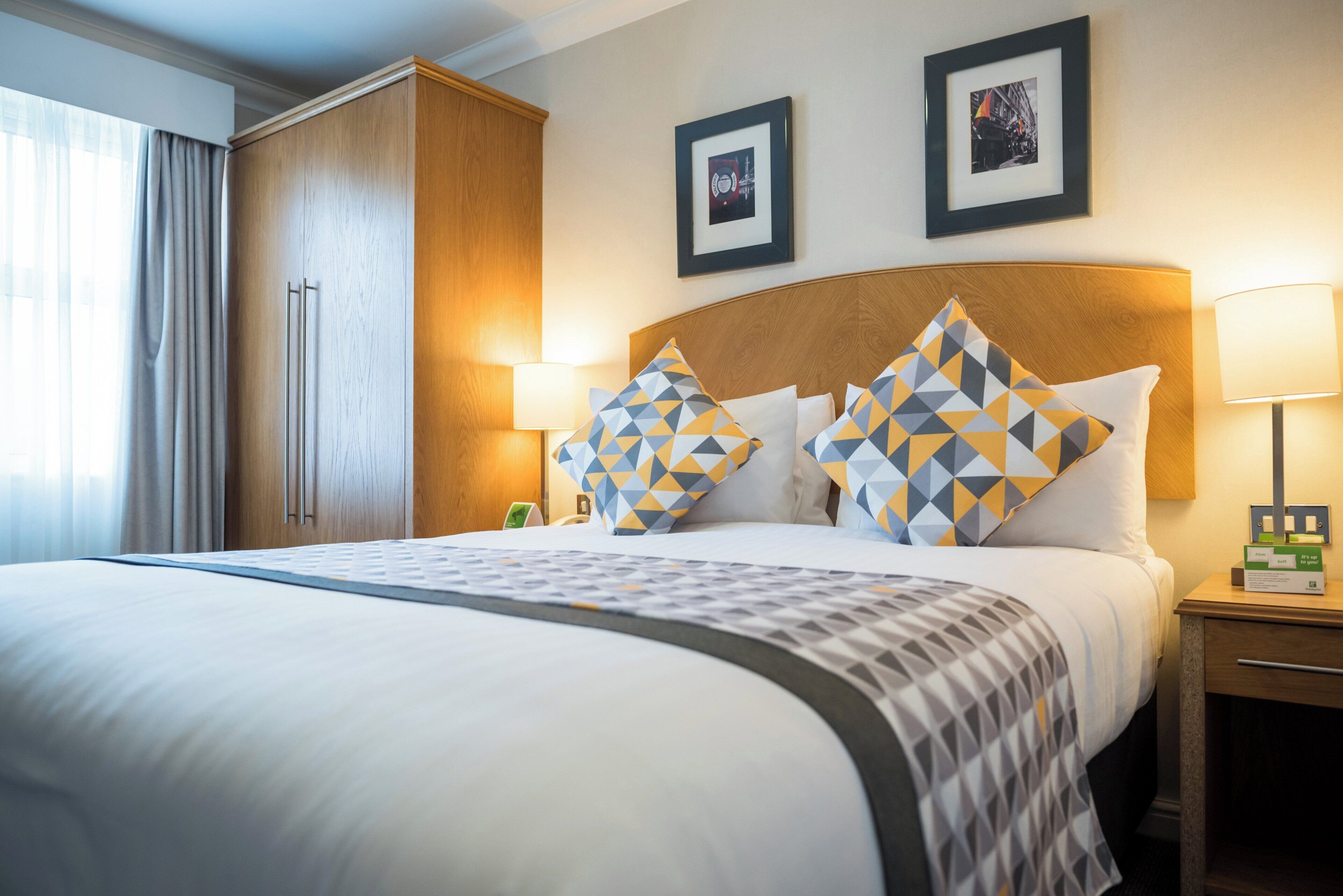 A hotel room with a large bed, two framed pictures on the wall, a wardrobe, and a nightstand with a lamp.