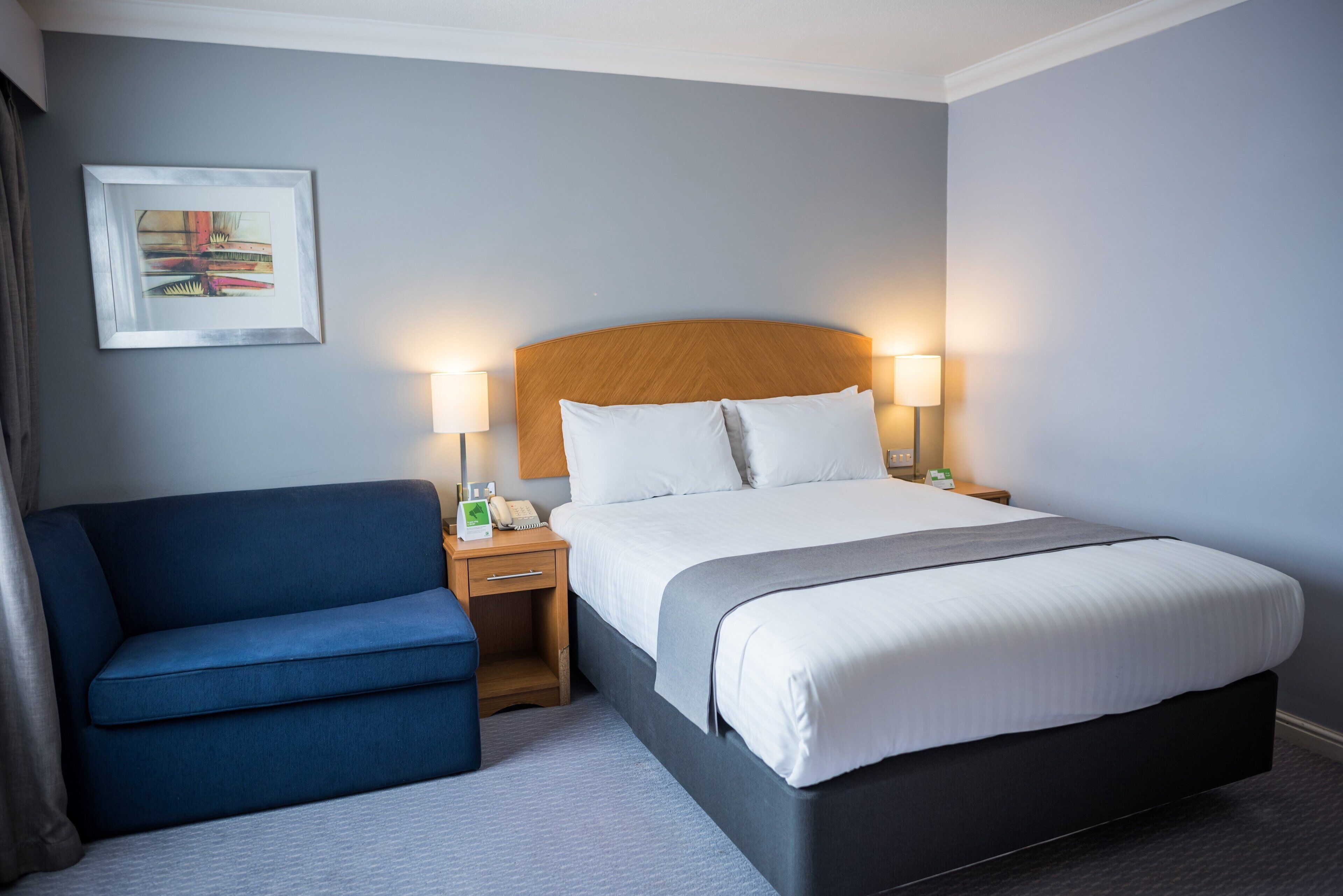 A hotel room with a bed, bedside lamps, a blue armchair, and a framed picture on the wall.