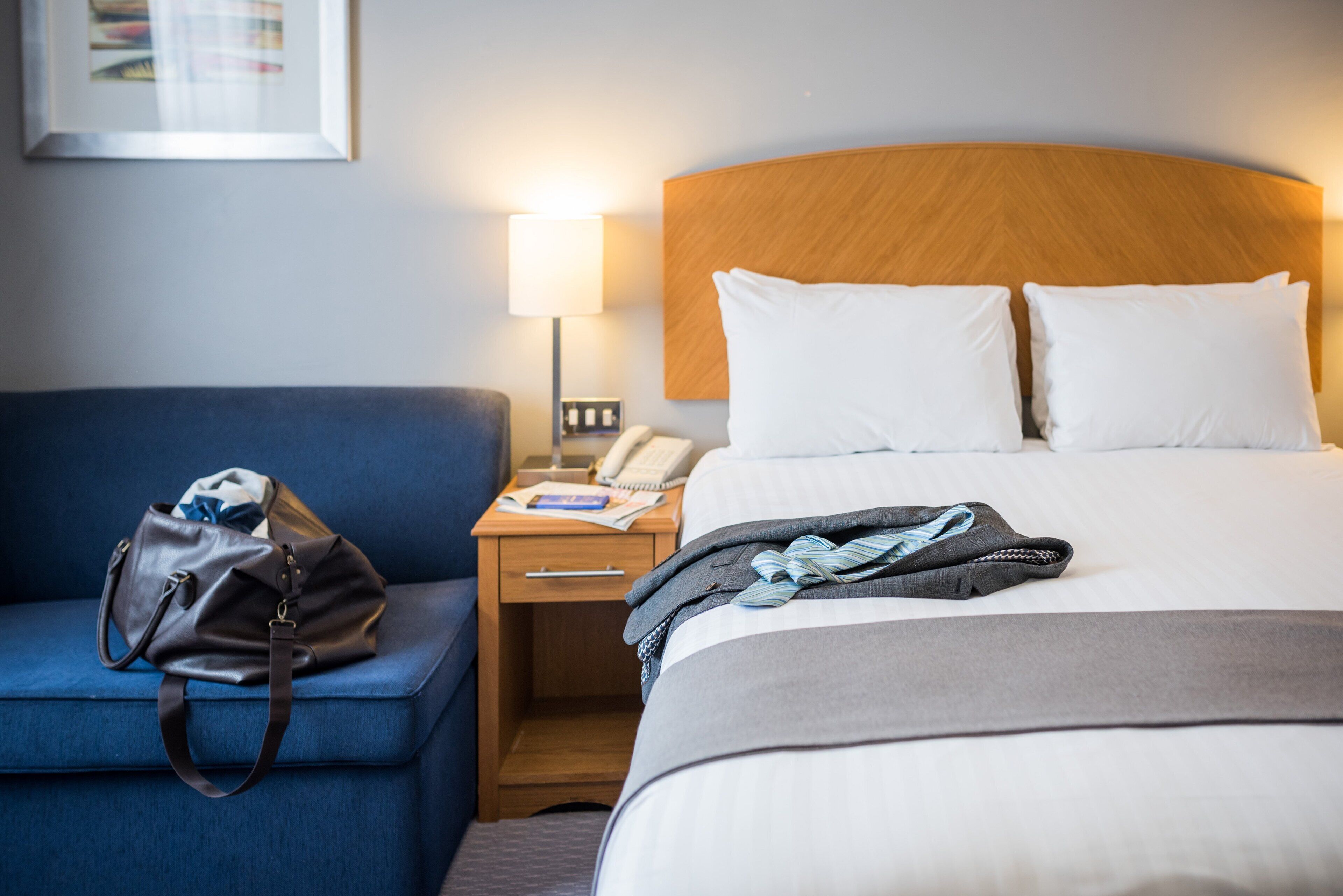 A hotel room with a bed, a nightstand, a lamp, a blue chair, and a handbag.
