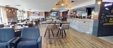 Un restaurante moderno con varias zonas de estar, incluyendo mesas y sillas, y una barra con una pantalla de menú.