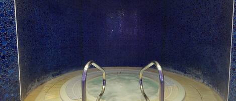 A circular hot tub with metal railings, surrounded by a tiled floor and walls with a blue mosaic pattern.