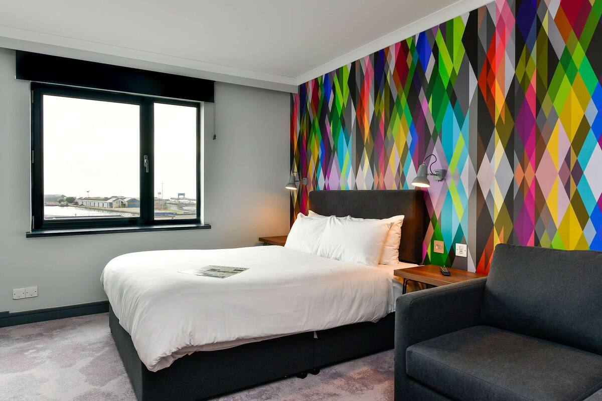 A modern hotel room with a large bed, a sofa, a colorful geometric wall, and a window with a view.