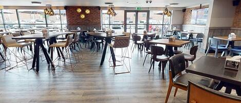 A spacious restaurant with multiple tables and chairs, large windows, and a brick wall.