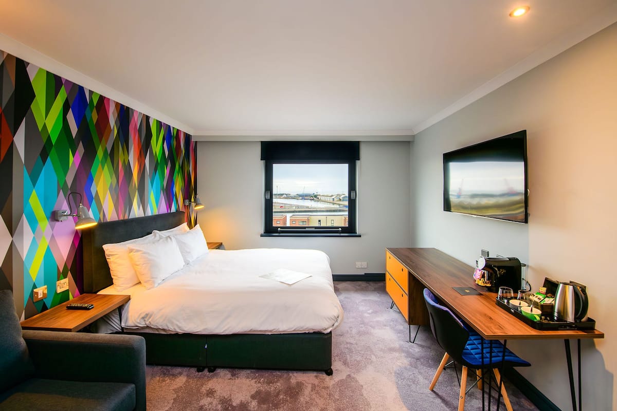 A modern hotel room with a large bed, a desk, and a view of the city through a window.