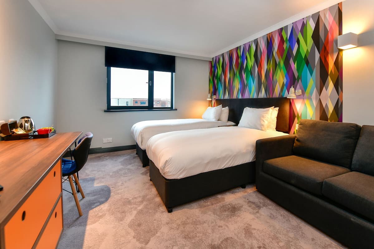 A hotel room with two beds, a desk, a chair, a sofa, and a colorful wall.