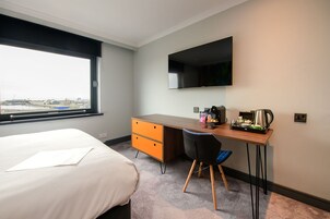 A hotel room with a bed, a desk with a chair, a flat-screen TV, and a window with a view of buildings.
