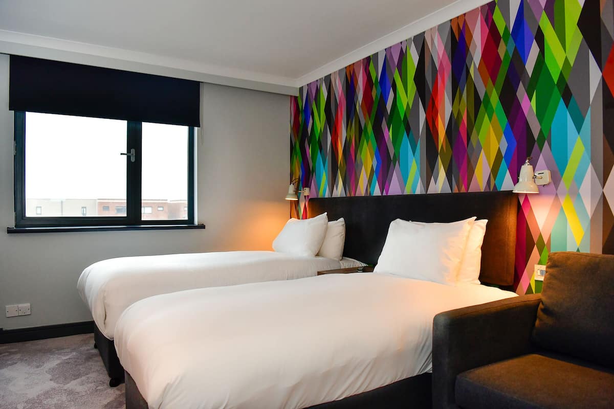A hotel room with two beds, a colorful geometric wall design, a window with a view, and a dark-colored armchair.