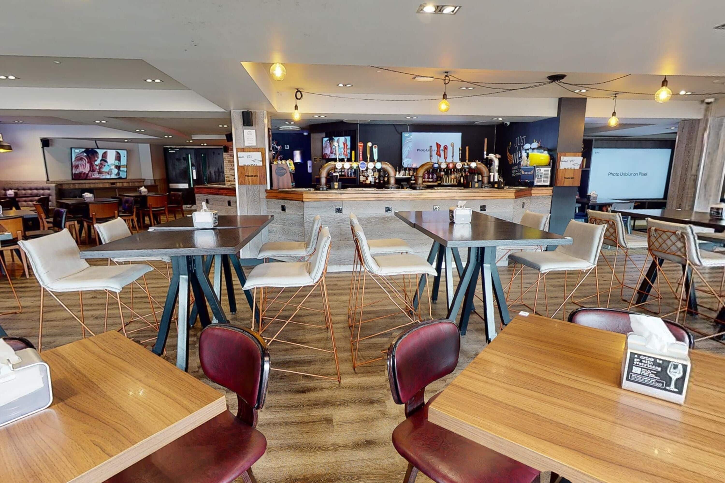 A modern restaurant with a bar area, multiple seating options including tables and chairs, and a television screen in the background.