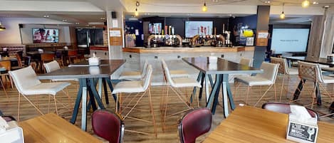 Un restaurante moderno con barra, diversas opciones de asientos, incluyendo mesas y sillas, y una pantalla de televisión al fondo.