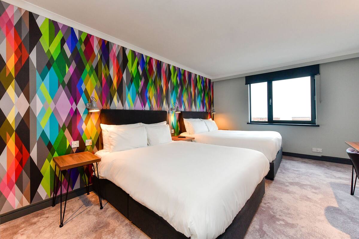 A hotel room with two beds, a colorful geometric-patterned wall, a window, and a small table.