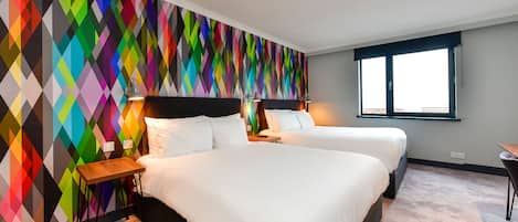 Habitación de hotel con dos camas, una pared de diseño geométrico y colorida, una ventana y una mesita pequeña.