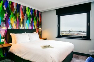 Ett hotellrum med en säng, en färgglad vägg med geometriskt mönster, ett fönster med utsikt över ett industriområde och ett nattduksbord med en lampa.