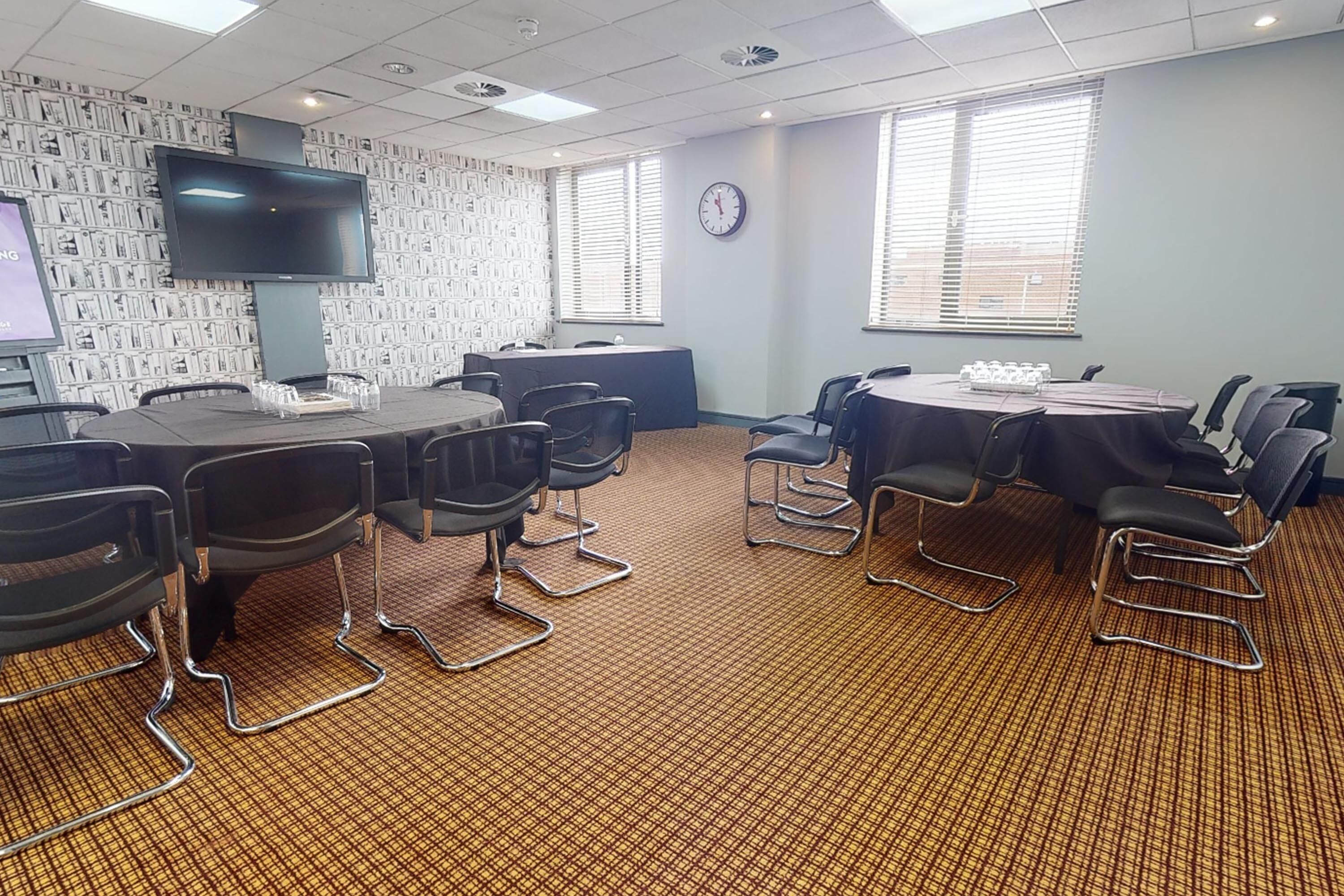 A conference room with a large flat-screen TV, multiple tables with chairs, and a patterned carpet.