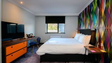 A modern hotel room with a bed, a desk with a computer, a television, and a colorful wall.