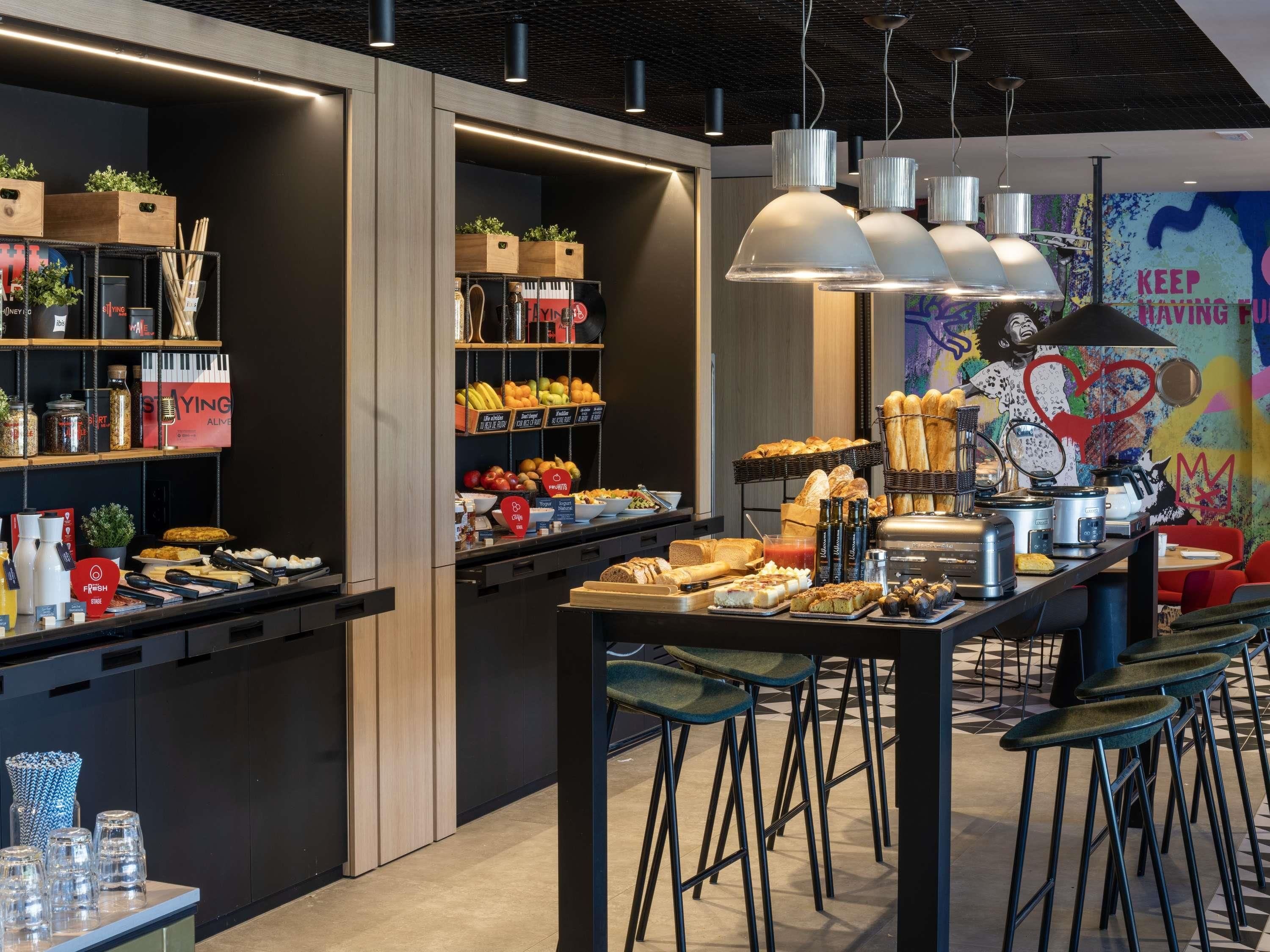 A modern café with a central counter displaying food and drinks, surrounded by bar stools and a colorful mural.