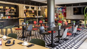 A modern cafe with a black and white checkered floor, multiple seating areas with black and red chairs, a bar area with shelves stocked with various items, and a large flat-screen TV mounted on the wall. - ibis Barcelona Aeropuerto Viladecans (Viladecans)