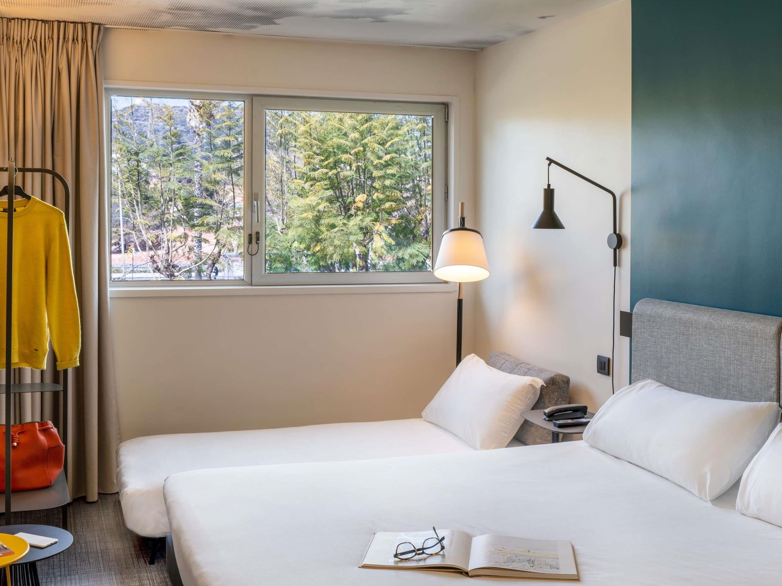 Habitación de hotel con cama, mesita de noche, lámpara y ventana con vista a árboles.