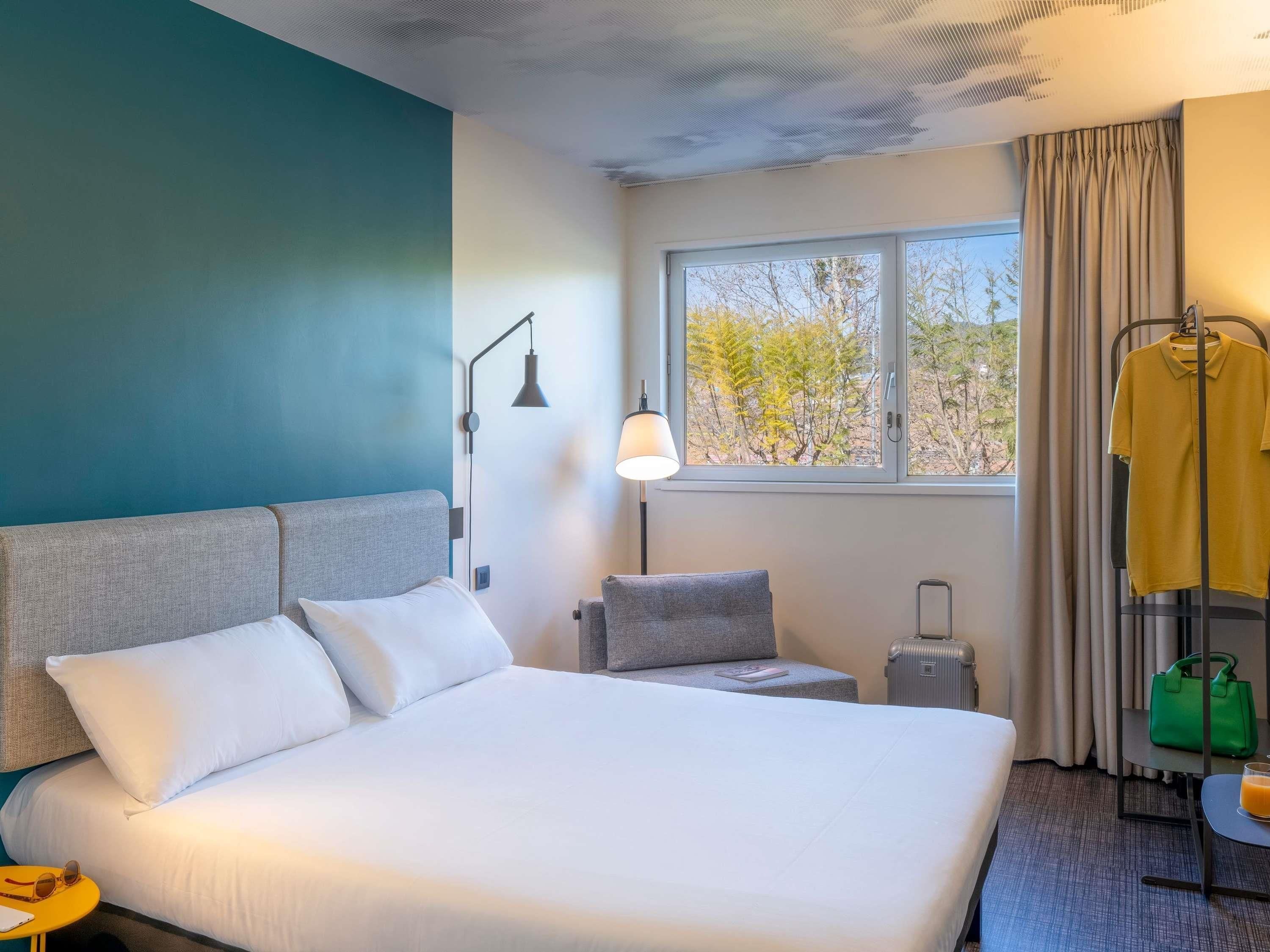 Een hotelkamer met een bed, een bureau, een stoel, een raam met uitzicht, een lamp en een kledingroede.