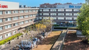 A multi-story hotel with a flat facade, a parking area with cars, and a garden area with trees and shrubs. - ibis Barcelona Aeropuerto Viladecans (Viladecans)