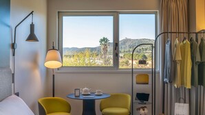 A hotel room with a bed, two yellow chairs, a table with a teapot and cups, a lamp, a window with a view of hills, and a clothing rack with hanging clothes. - ibis Barcelona Aeropuerto Viladecans (Viladecans)