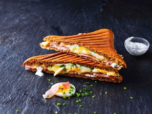 A sandwich cut in half, placed on a dark surface with a small bowl of sauce nearby. - ibis Barcelona Aeropuerto Viladecans (Viladecans)
