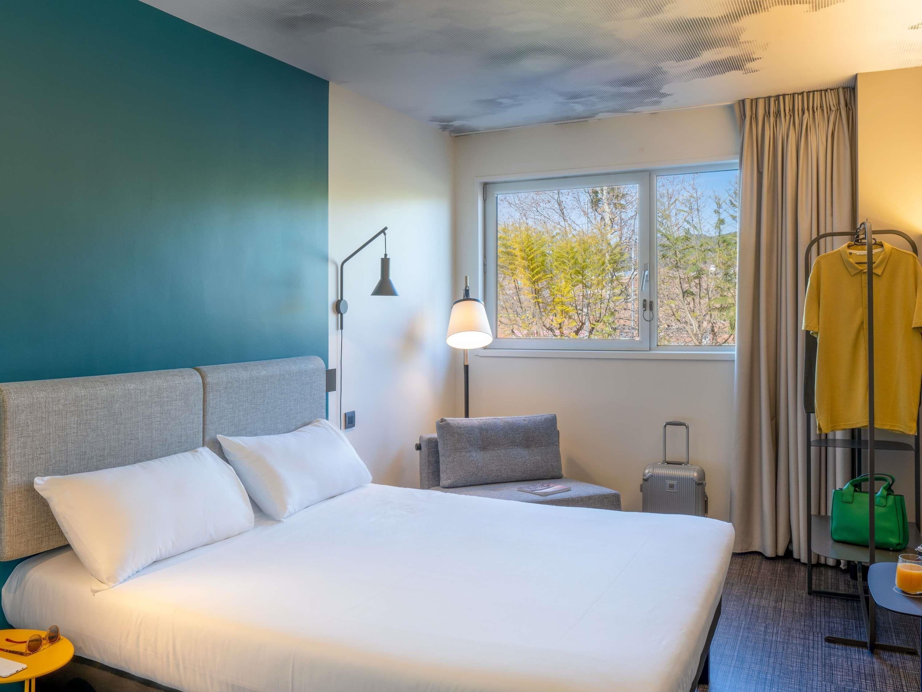 A hotel room with a bed, a window with a view of trees, a lamp, a chair, a small table with a glass of orange juice, and a coat rack with a yellow shirt and a green bag.