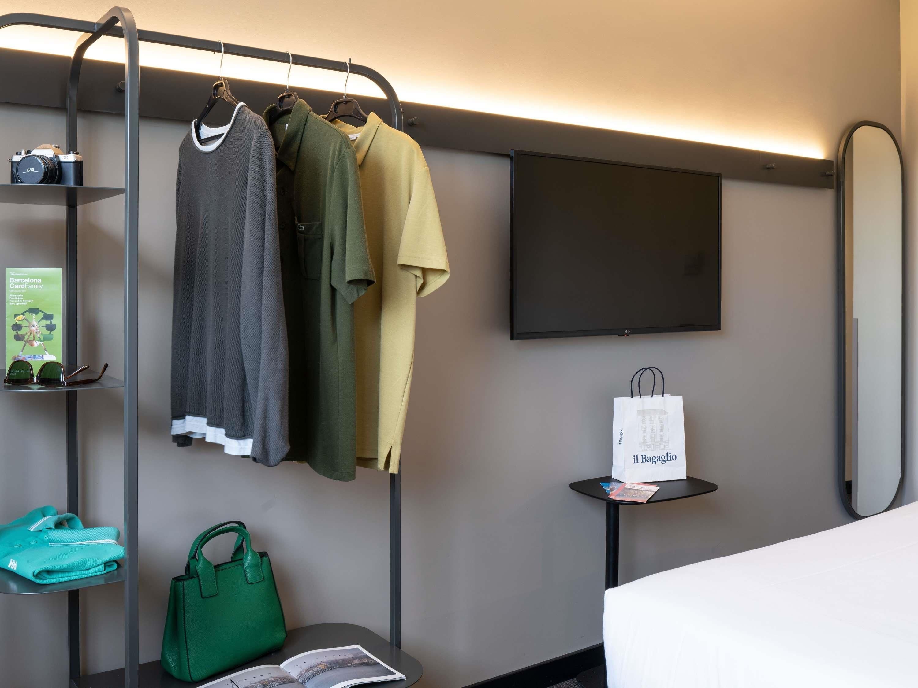 Habitación de hotel con perchero, televisor de pantalla plana, una mesita con una bolsa de compras, un bolso verde y una revista.