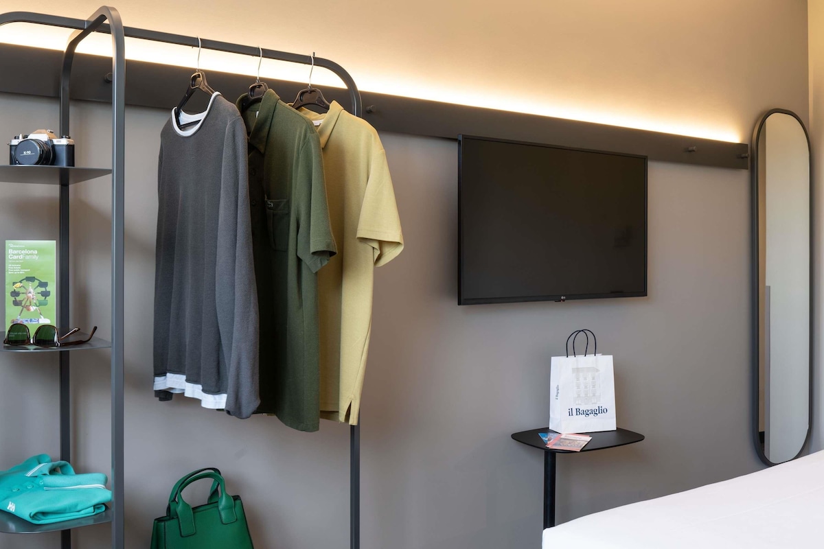 A hotel room with a clothing rack, a flat-screen TV, a small table with a shopping bag, a green handbag, and a magazine.