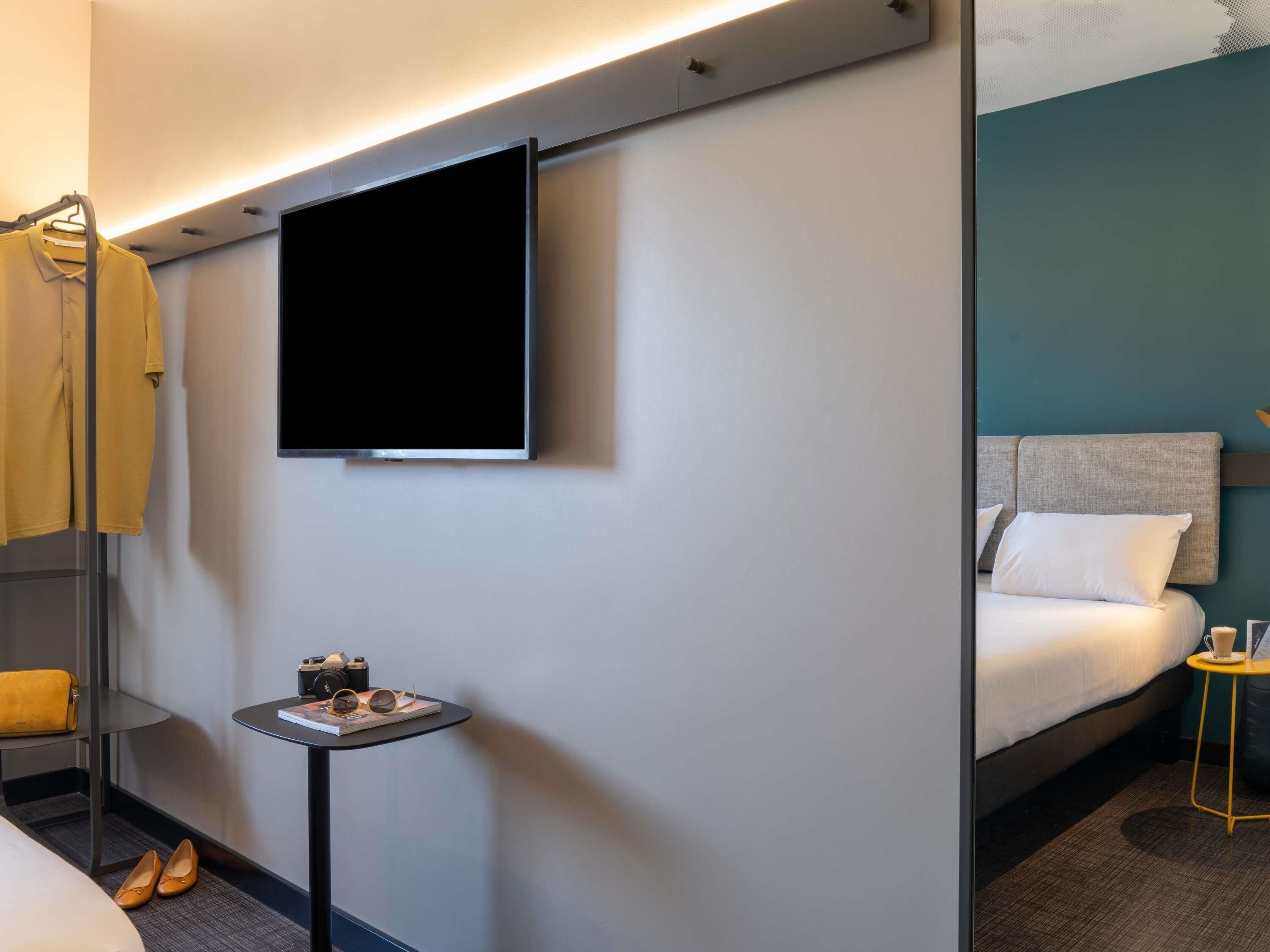 Habitación de hotel con una cama, una mesita con una taza y un libro, un televisor montado en la pared y un abrigo colgado en un perchero.