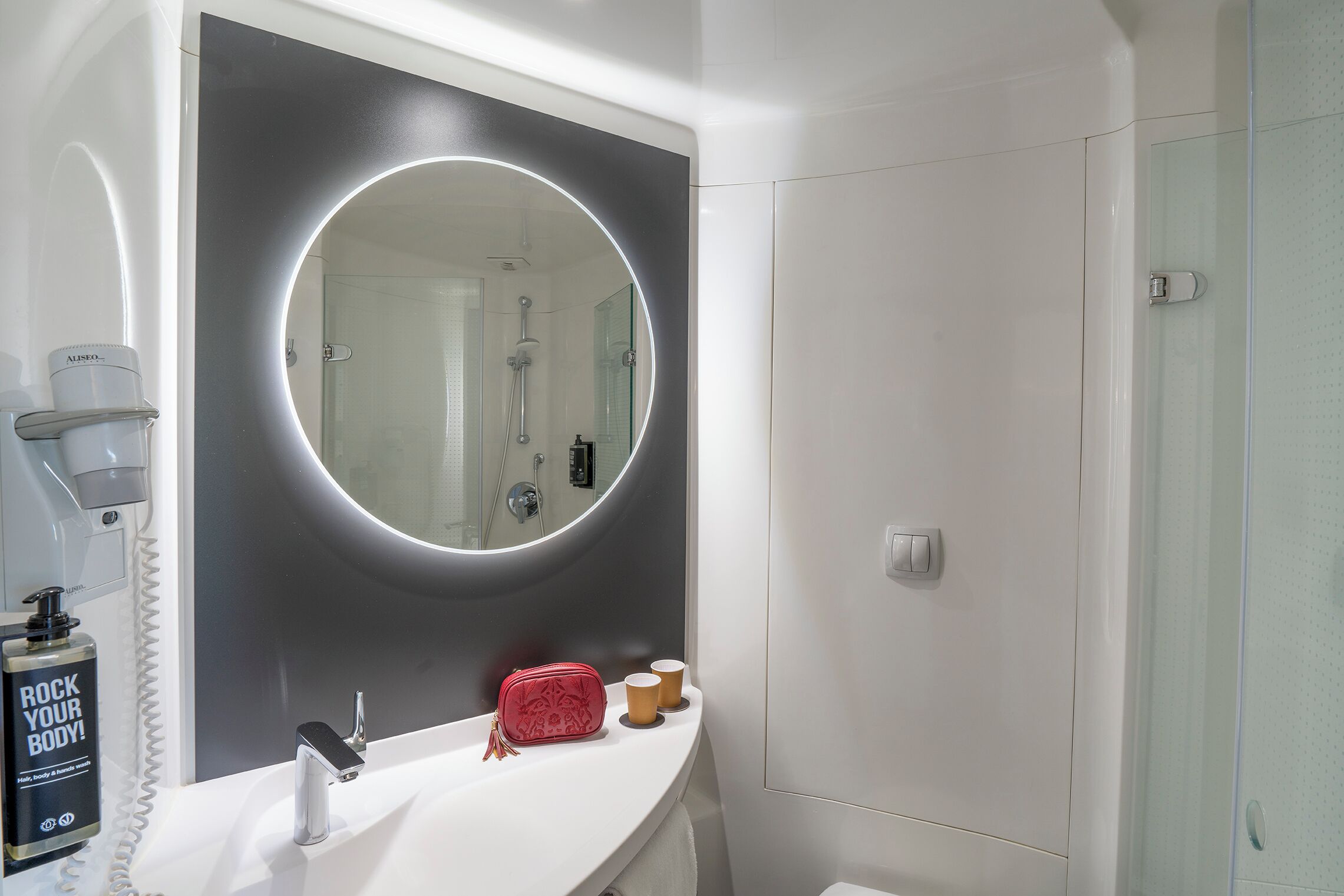 Un baño con un espejo grande, un lavamanos blanco, un cabezal de ducha manual y una botella de champú.