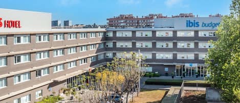 Un hotel de varios pisos con fachada plana, un estacionamiento con coches y una zona ajardinada con árboles y arbustos.