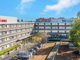 Um hotel com vários andares, fachada plana, área de estacionamento com carros e um jardim com árvores e arbustos.