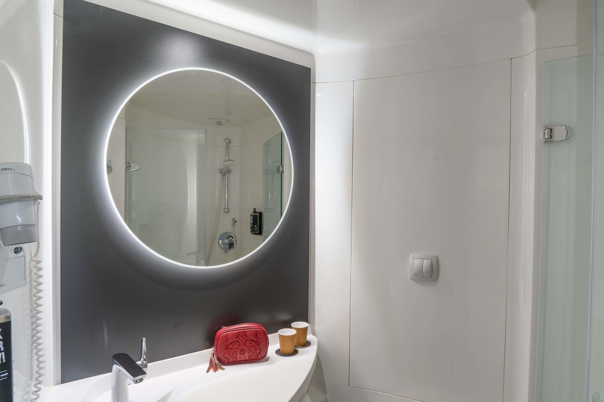 Un baño con un espejo redondo, un lavamanos blanco, un bolso rojo y una cabina de ducha de vidrio.