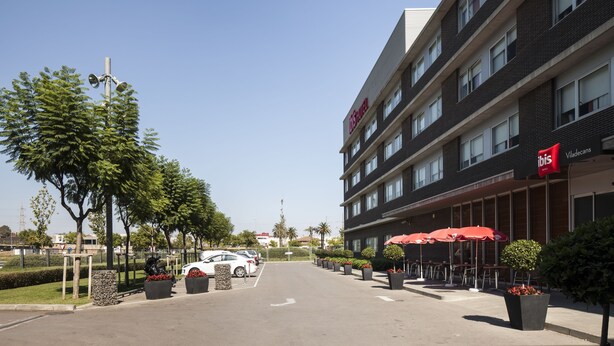 A hotel with a modern design, featuring a parking lot, outdoor seating area, and a clear sky. - ibis Barcelona Aeropuerto Viladecans (Viladecans)