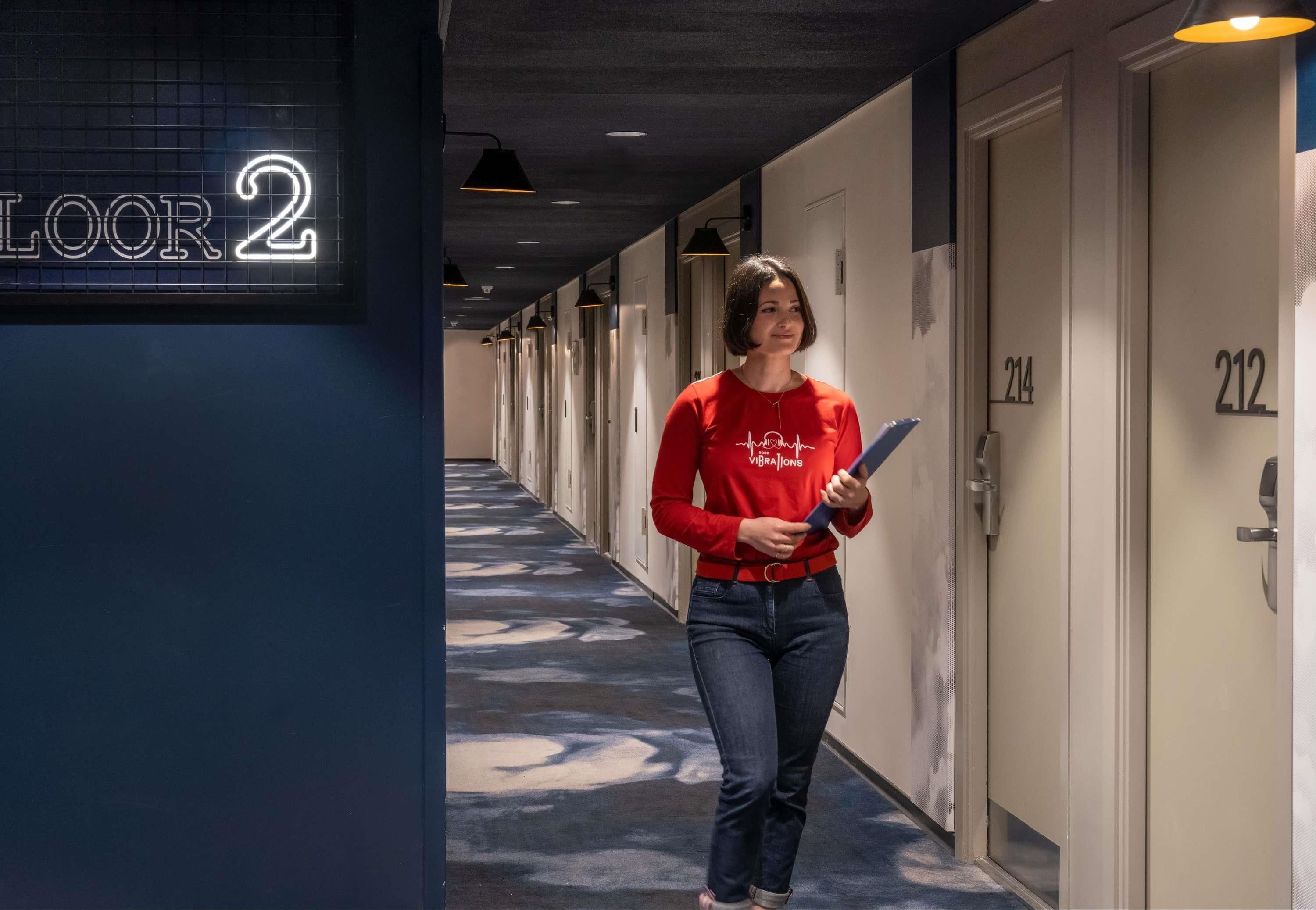 Pasillo de hotel con puertas numeradas 214 y 212, un letrero de neón que indica 'Segundo piso' y una persona sosteniendo una tableta.