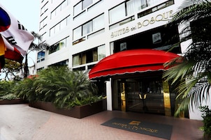 Entrada de un hotel con toldo rojo, palmeras y un letrero que dice "SUITES del BOSQUE".