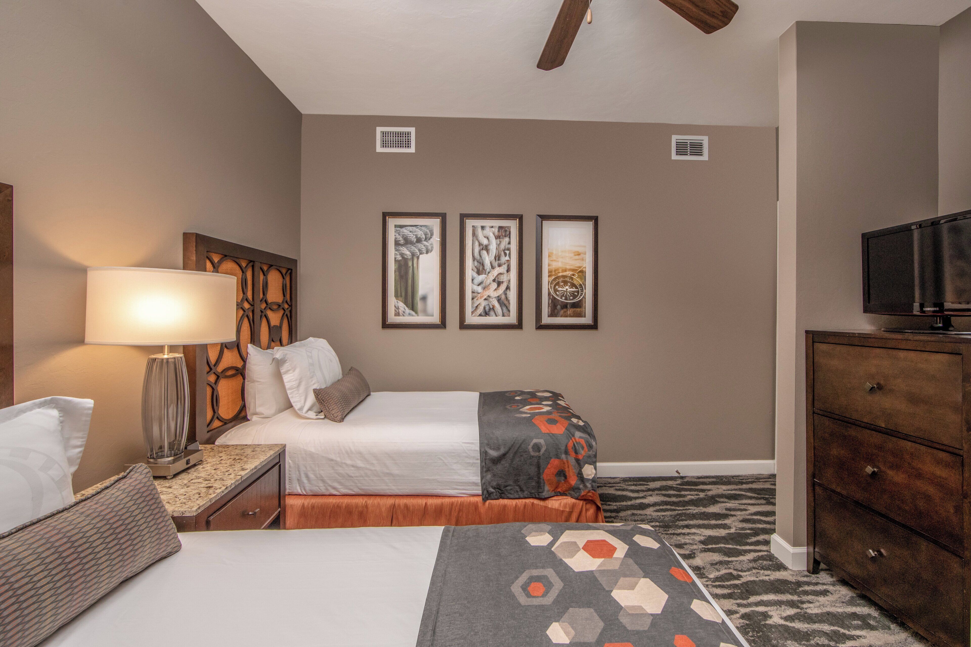 A hotel room with two beds, a desk with a lamp, a dresser, and three framed pictures on the wall.