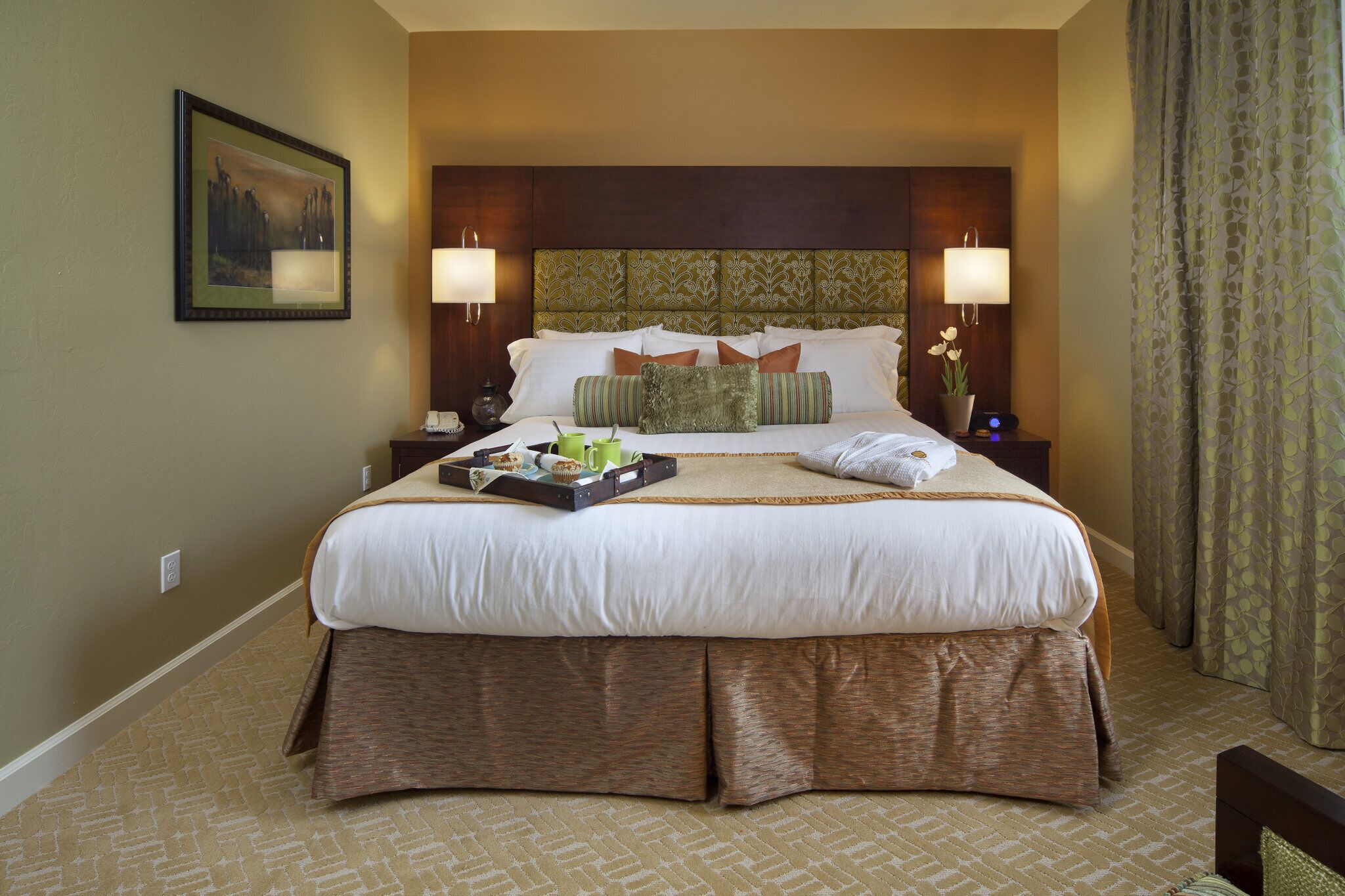 A hotel room with a large bed, two bedside tables, a headboard with a patterned design, a framed picture on the wall, and a window with curtains.