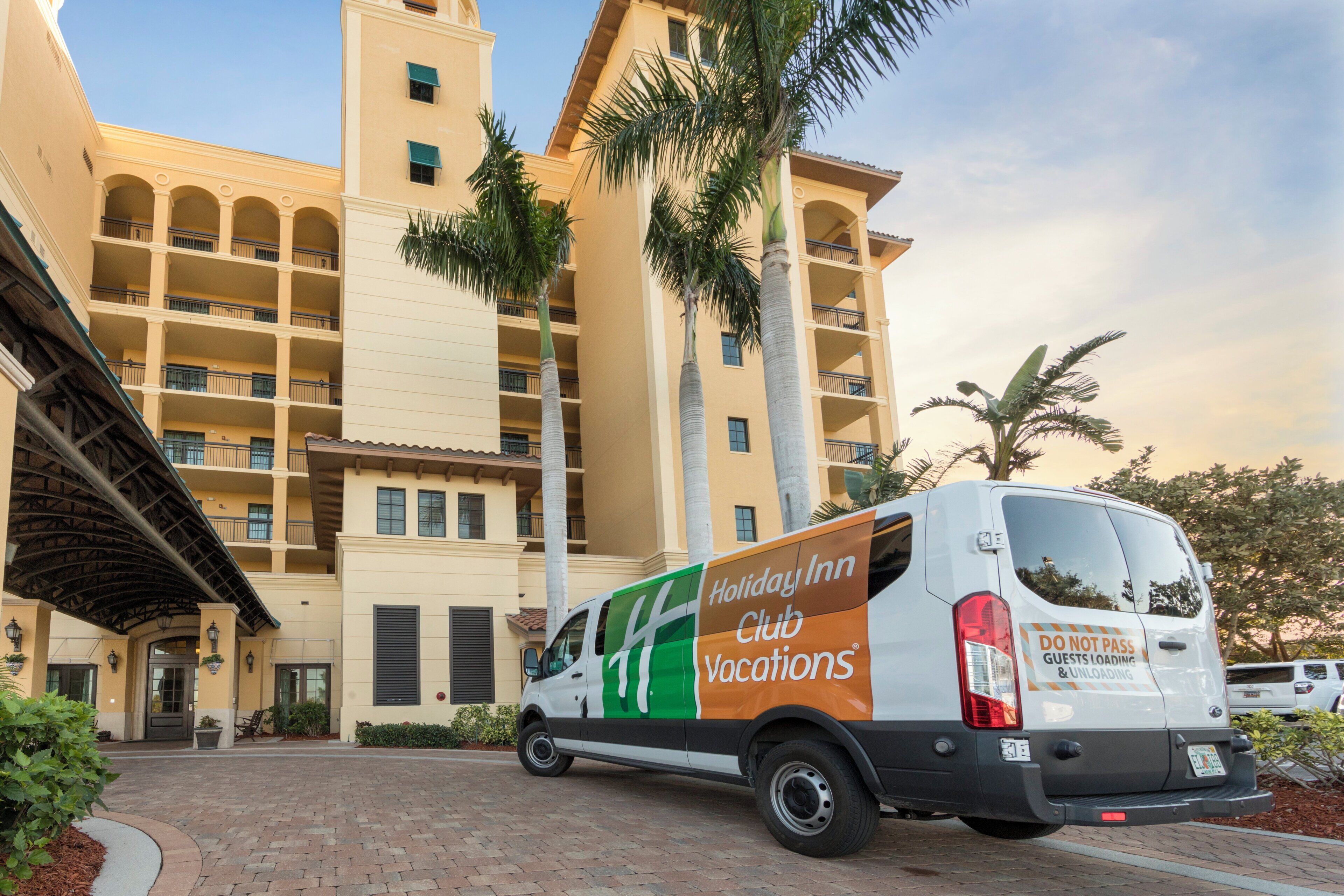 Een Holiday Inn Club Vacations busje geparkeerd voor een meerlaags gebouw met balkons en palmbomen.