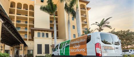 Een Holiday Inn Club Vacations busje geparkeerd voor een meerlaags gebouw met balkons en palmbomen.