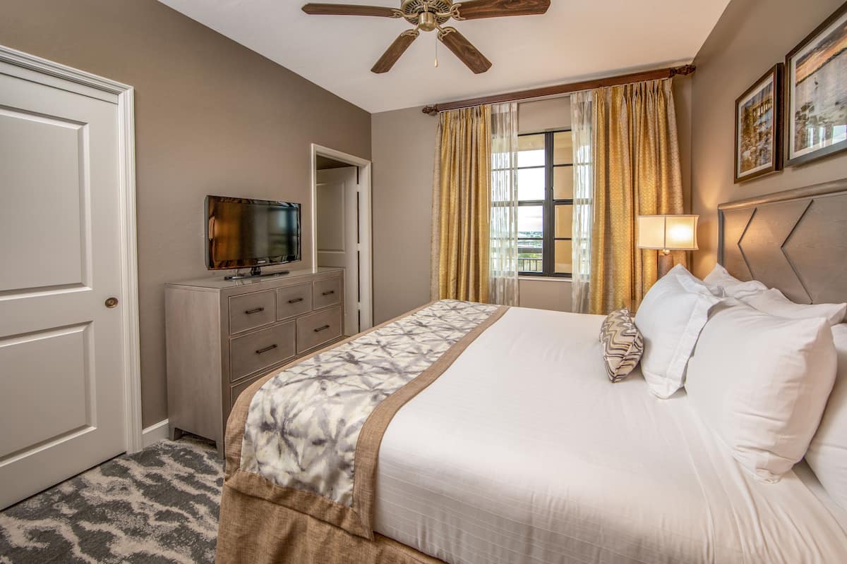 A hotel room with a bed, a television, a wardrobe, a ceiling fan, and a window with curtains.