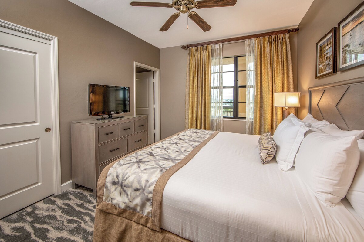 A hotel room with a bed, a television, a ceiling fan, a window with curtains, and a door.