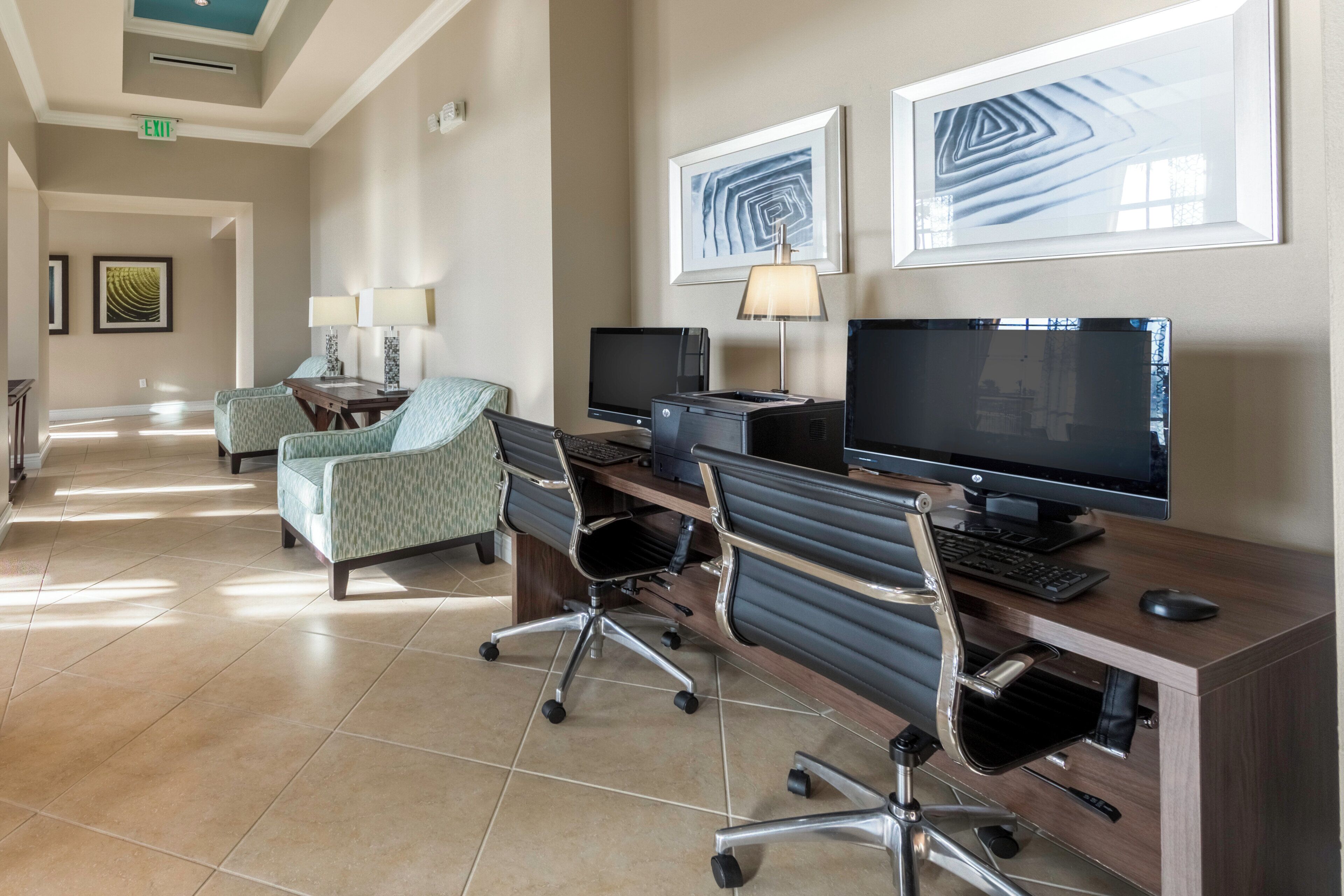 A hotel lobby with a reception desk, two computer workstations, a seating area with chairs, and artwork on the walls.