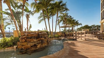A resort pool area with palm trees, a stone waterfall, and outdoor seating.