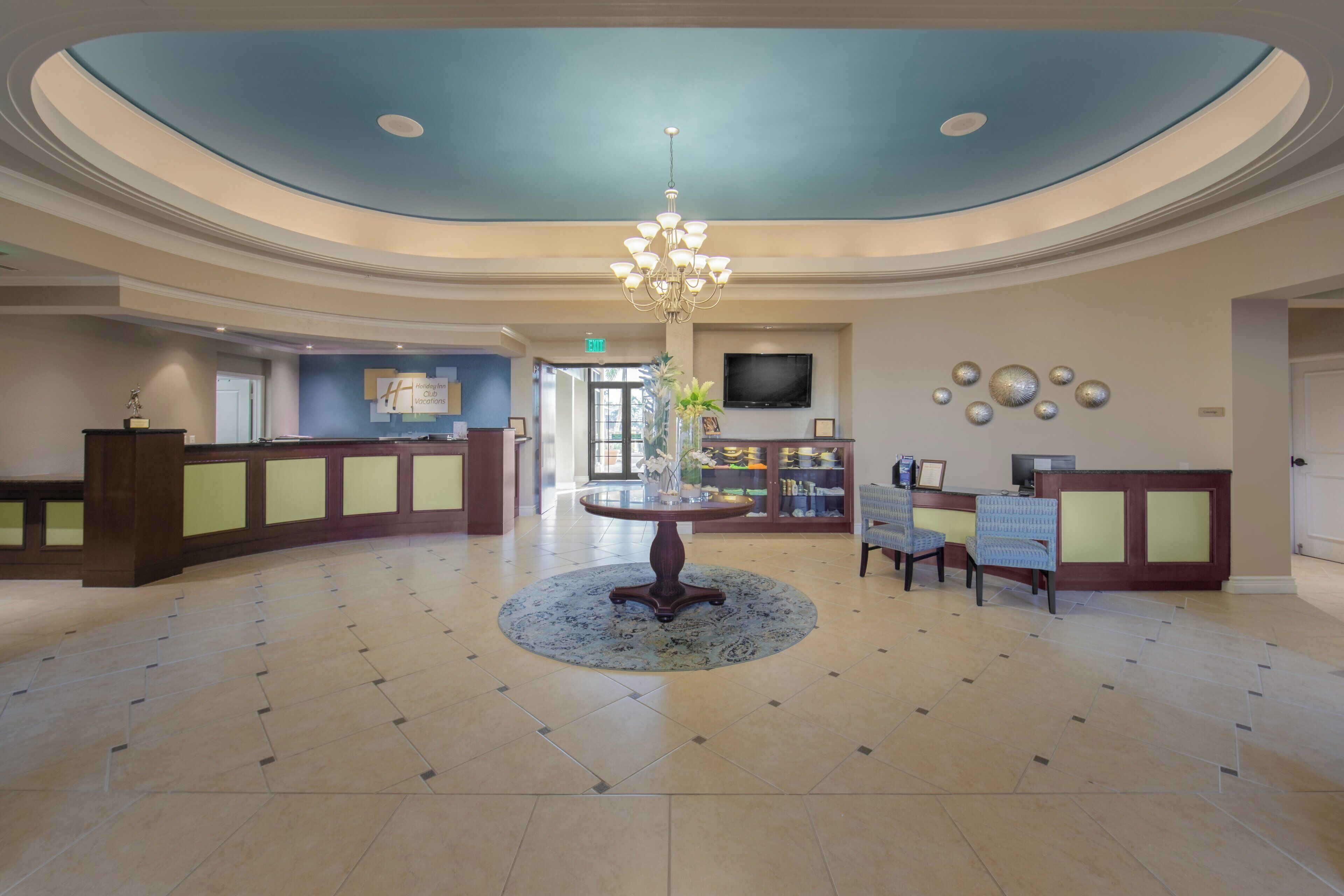 A hotel lobby with a reception desk, a chandelier, a dining area with chairs, and a television on the wall.