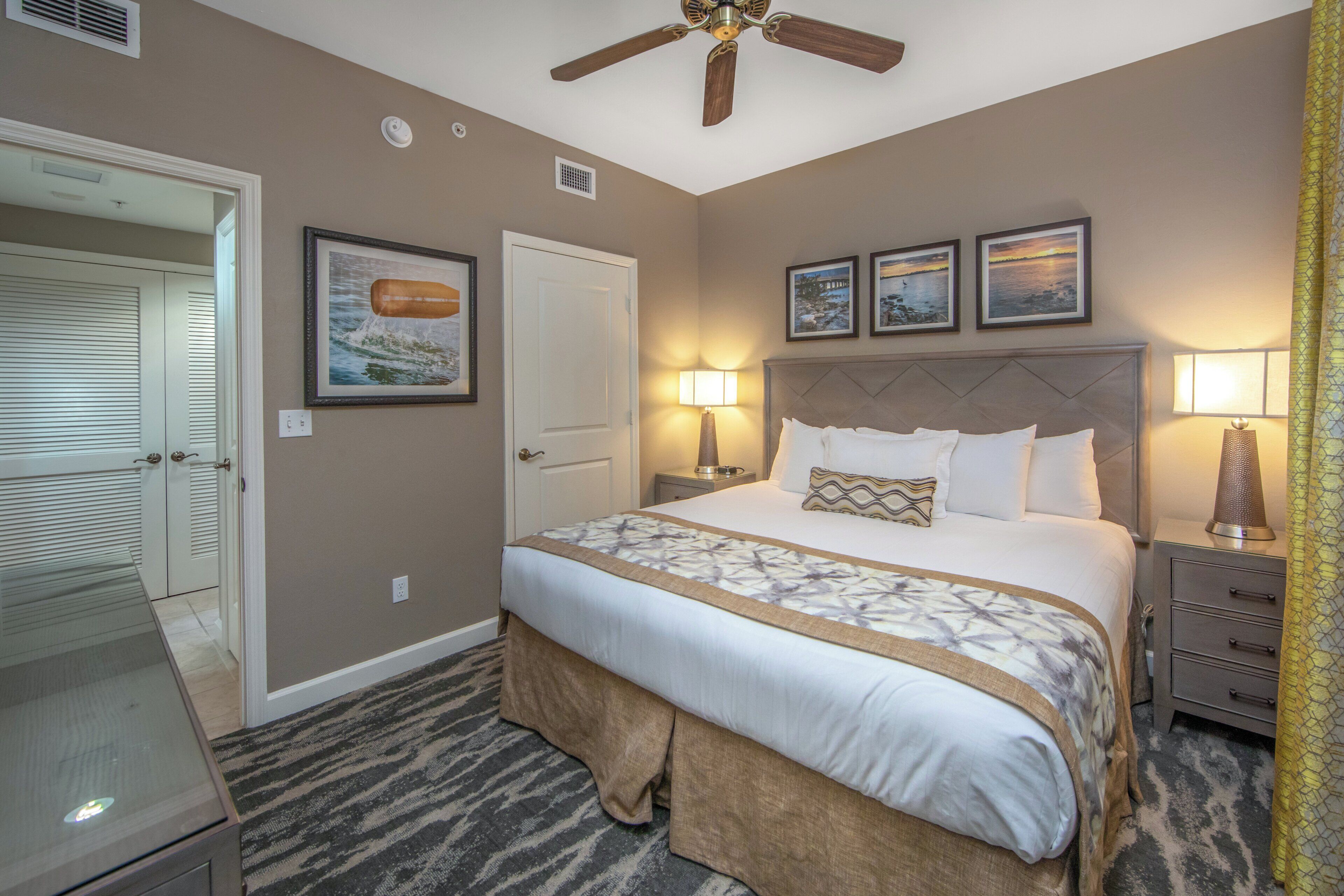 A hotel room with a bed, bedside lamps, a ceiling fan, a door, and three framed pictures on the wall.