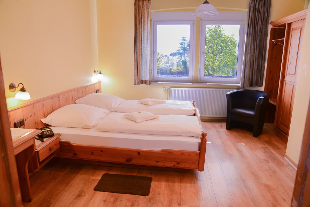 A neatly made bed with white linens, a wooden nightstand with a telephone, a chair, and a window with curtains.
