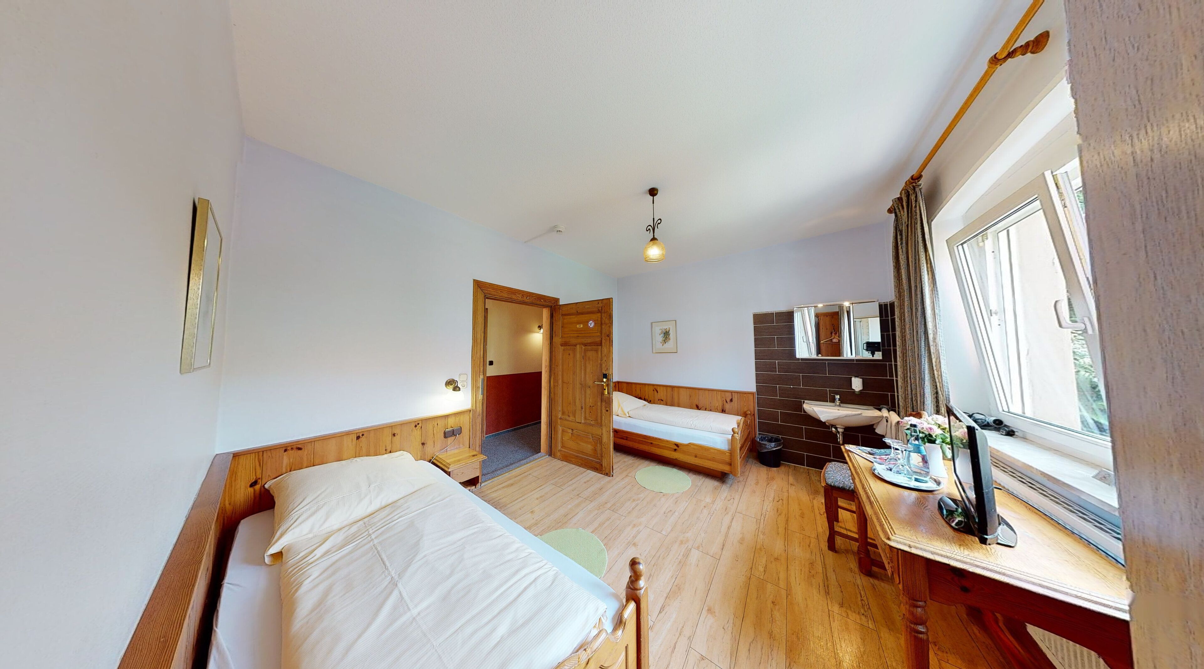 A room with two beds, a wooden floor, a small table with a mirror, and a window with curtains.