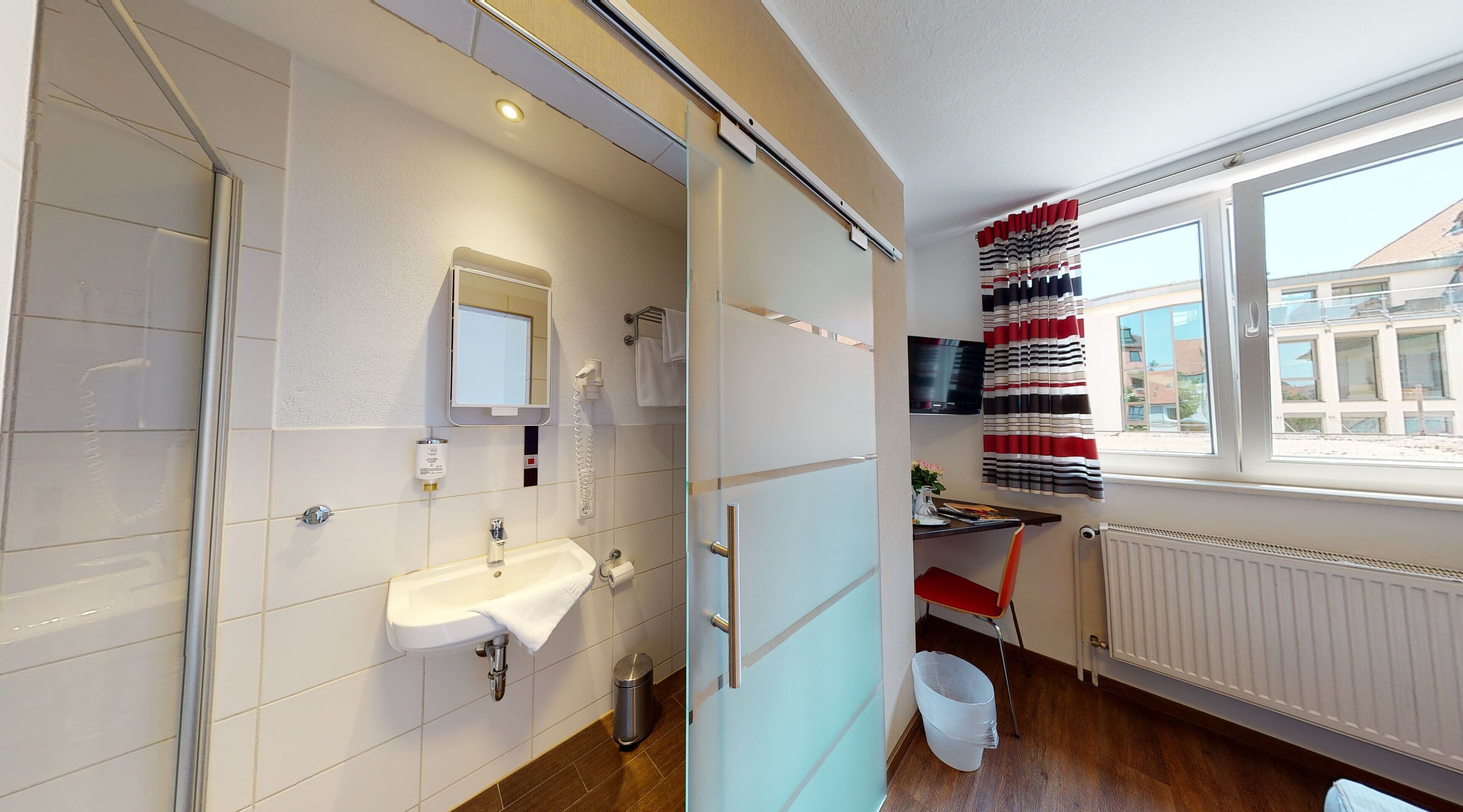 Un baño moderno con lavamanos, espejo y retrete. Se ve una ventana con vistas a edificios.